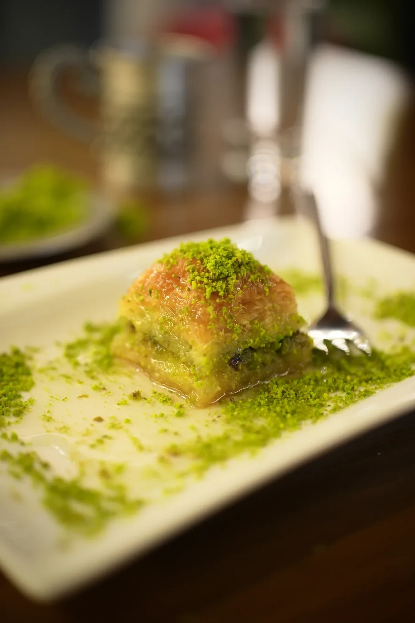 Einzelnes Stück Pistazien-Baklava mit Gabel auf weißem Teller, bestreut mit gemahlenen Pistazien